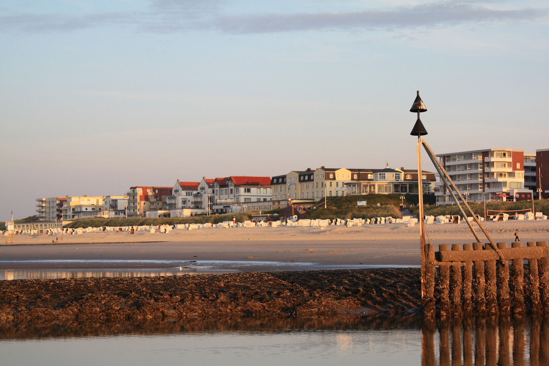 Blick vom Meer aus über den Sandstrand mit weit entfernten Strandkörben auf Häuser der Strandpromenade, im Vordergrung ist Wasser und ein Seezeichen