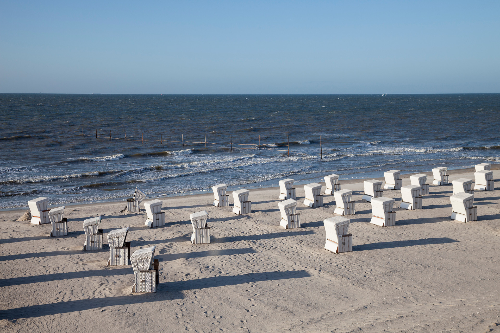 Blick über den Sandstrand aufs Meer, vor dem Wasser stehen viele weiße Strandkörbe, die alle zur Sonne ausgerichtet sind