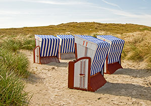 Vier blau-weiß gestreifte Strandkörbe stehen auf Sandboden in Mitten der Dünen, der Himmel ist blau.