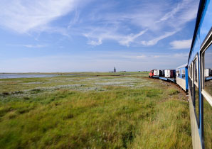 Ein Blick aus dem Zugfenster: Man sieht grüne Wiesen, viel Himmel und einen Teil des Zuges selbst. Am Horizont in Bildmitte ragt der Westturm von Wangerooge.